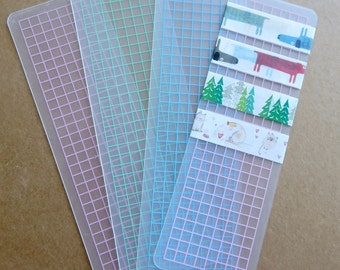May include: Four clear, rectangular bookmarks with a grid pattern in pink, green, and blue. One bookmark has decorative tape with dog, cat, and tree designs. The bookmarks are made of a translucent material.
