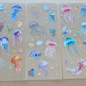 May include: Three sheets of decorative stickers featuring various jellyfish designs in shades of blue, purple, orange, and pink. The stickers also include seashells, coral, and star accents, all on a transparent background.