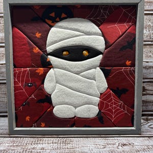 May include: A square wall art piece featuring a fabric-pieced mummy character in white bandages with glowing orange eyes, centered against a dark red background patterned with black bats, spiderwebs, and orange jack-o'-lanterns in a gray frame.