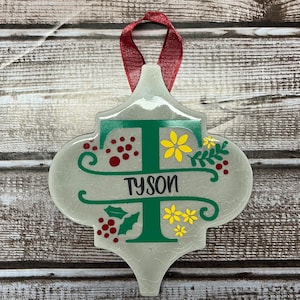 May include: A white ceramic Christmas ornament shaped like a stylized tree with a green letter T and the name Tyson written in black. The ornament is decorated with red berries, green leaves, and yellow flowers.