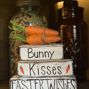 May include: Three wooden blocks stacked on top of each other. The top block says "Bunny", the middle block says "Kisses" with a small carrot image, and the bottom block says "Easter Wishes". Two orange carrots are tied together with twine and placed on top of the blocks.