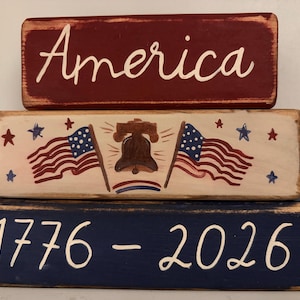 May include: A patriotic wooden block set with three stacked rectangular blocks. The top block is burgundy with the word "America" in white script. The middle block features American flags, stars, and a bell. The bottom block is navy blue with the numbers "1776 - 2026".
