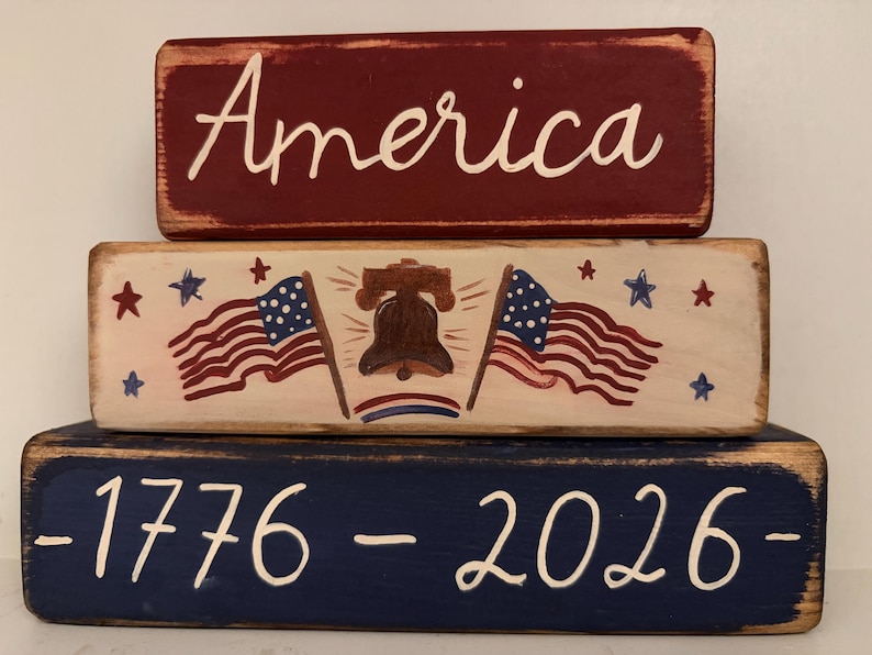 May include: A patriotic wooden block set with three stacked rectangular blocks. The top block is burgundy with the word "America" in white script. The middle block features American flags, stars, and a bell. The bottom block is navy blue with the numbers "1776 - 2026".