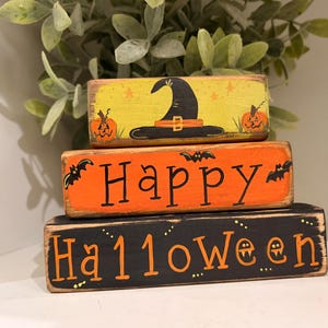 May include: Three stacked wooden blocks with Halloween-themed designs. The top block is yellow with a witch hat and pumpkins. The middle block says "Happy" in orange. The bottom block says "Halloween" in orange on black.