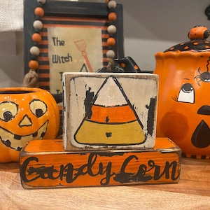 May include: Two wooden signs with Halloween themes. The top sign is white with black lettering and a painted image of a candy corn. The bottom sign is orange with black lettering that reads "Candy Corn".
