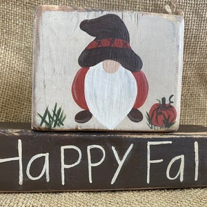 May include: A wooden block sign with a painted gnome and pumpkin, stacked on top of a brown wooden block sign with the text "HAPPY FALL" in white paint.