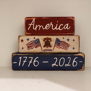May include: Three stacked wooden blocks with patriotic themes. The top block is red with the word "America" in white script. The middle block has American flags, stars, and a bell. The bottom block is blue with "-1776-2026-" in white.