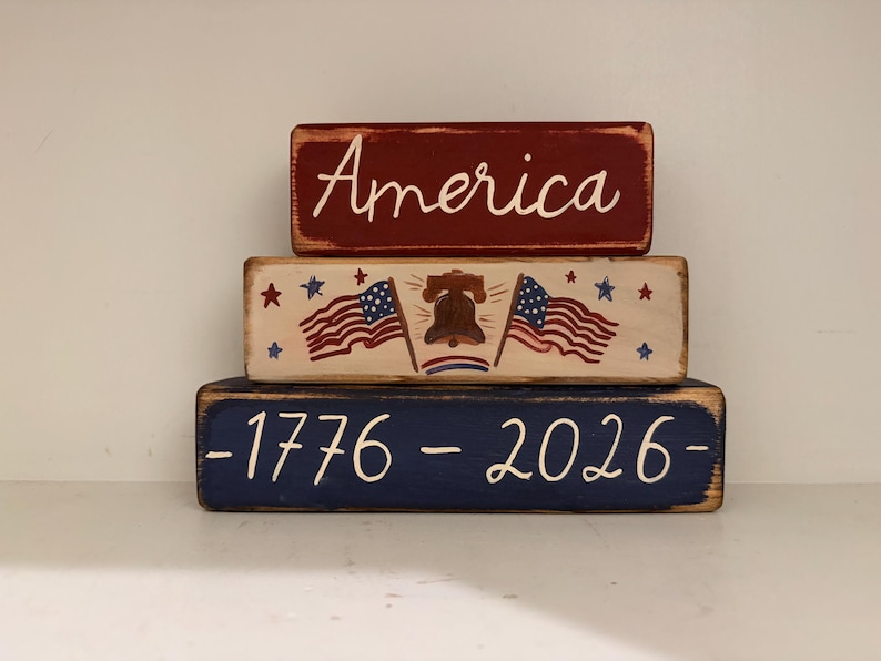 May include: Three stacked wooden blocks with patriotic themes. The top block is red with the word "America" in white script. The middle block has American flags, stars, and a bell. The bottom block is blue with "-1776-2026-" in white.