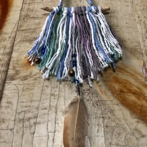 May include: A handmade wall hanging with a wooden dowel, blue, white, and green yarn, and a brown feather. The yarn is woven into a fringe and has beads and bells hanging from it.