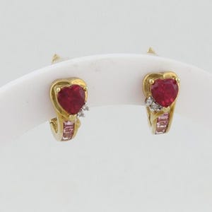 Peut inclure: Une paire de boucles d'oreilles en or, chacune ornée d'une pierre rouge en forme de cœur. De petits pierres blanches et roses rectangulaires ornent le design. Les boucles d'oreilles sont présentées sur un fond blanc.