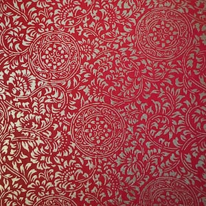 May include: A red background with a repeating pattern of silver floral and circular designs. The intricate pattern features stylized flowers and swirling leaves, creating a rich, textured appearance. The design is symmetrical and detailed.