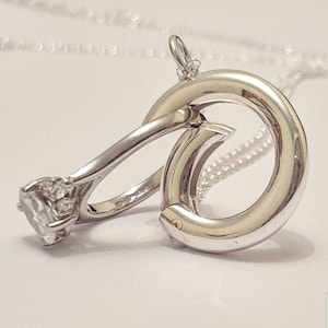 May include: A silver ring with a large, clear gemstone and smaller accent stones is attached to a silver chain. The ring is designed to be worn as a pendant.