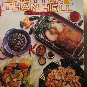 May include: A cookbook titled "Jane Butel's Hotter Than Hell: Revised Edition" with a photo of a variety of spicy dishes, including a roasted chicken, a salad with tomatoes and lettuce, and a plate of shrimp.