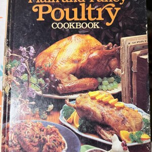 以下が含まれることがあります： 「The Southern Heritage Plain and Fancy Poultry Cookbook」というタイトルのヴィンテージ料理本。表紙には、ローストターキー、アヒル、その他の鶏肉料理、ブドウ、オレンジのスライスが描かれています。この本は料理ガイドです。
