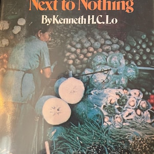 May include: A book titled "Chinese Cooking on Next to Nothing" by Kenneth H.C. Lo. The cover features a photo of a market stall with a variety of fresh produce, including vegetables, gourds, and potatoes.