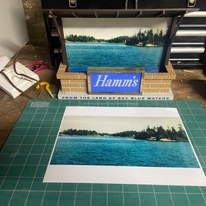 May include: A vintage Hamm's beer sign featuring a scenic lake view. The sign has a brick-like base and a blue illuminated panel with the Hamm's logo. The text "FROM THE LAND OF SKY BLUE WATERS" is also visible.