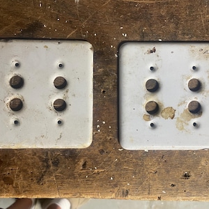 May include: Two vintage, square, off-white light switch covers. Each cover has two large circular holes for the switches and four smaller screw holes. The covers show signs of age and wear.