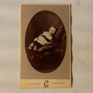 May include: Sepia-toned antique photograph of a child reclining in an ornate chair. The child wears a white dress with a dark stripe and dark shoes. The photograph is in an oval frame with the text "J. CHENHALL TAVISTOCK" at the bottom.
