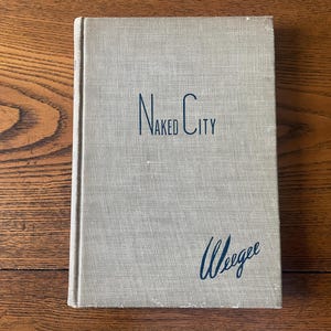 May include: A vintage book titled "Naked City" with the author's name, Weegee, in a cursive font. The book has a light gray cover with the title in a dark blue, stylized font. The book is lying on a wooden surface.