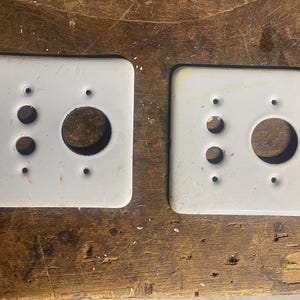 May include: Two vintage white electrical outlet covers. Each square cover has a large central hole and smaller holes for switches. The covers are slightly worn, showing signs of age and use. They sit on a wooden surface.