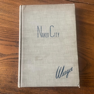 May include: A hardcover book titled "Naked City" with the author's name, Weegee, in cursive script. The book has a light tan cover with the title in a dark blue font. The book is resting on a wooden surface.