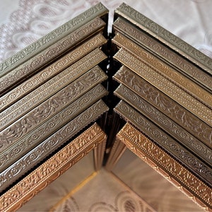 May include: A selection of decorative picture frames in gold, bronze, and silver tones. The frames display intricate embossed floral and geometric designs, giving a vintage feel. The frames are stacked at an angle, highlighting their decorative edges. Metric measurements.