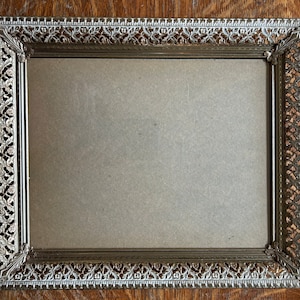 Puede incluir: Marco de fotos rectangular de metal vintage con detalles de filigrana ornamentados. El marco tiene un acabado dorado con detalles en blanco y un respaldo de color neutro. El marco está vacío y listo para una foto.