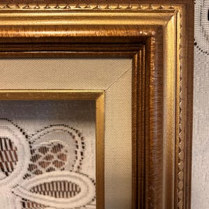 May include: A rectangular, ornate gold picture frame with a textured, warm-toned finish. The frame has a double border design and a neutral-colored fabric insert. The frame is empty.