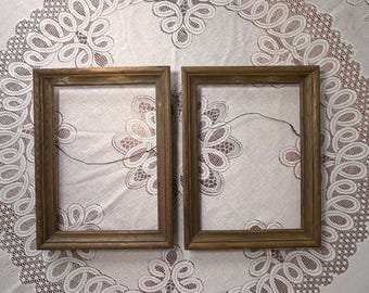 May include: Two rectangular, wooden picture frames with a warm, brown finish. The frames are empty, with a simple, classic design. They rest on a white tablecloth with an intricate, lace-like pattern. The frames are ready to display photos or artwork.