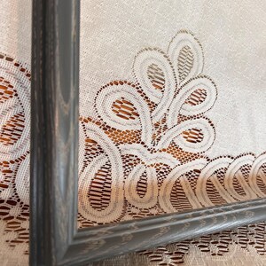 May include: A gray wooden picture frame with a distressed finish. The frame is angled, showing the corner and side profile. The background is a white lace tablecloth with a floral design.