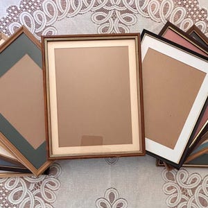 May include: A collection of rectangular picture frames arranged in a fan shape. The frames feature various wood tones and metallic finishes, each containing a mat board in colors including white, cream, gray, blue, and pink, surrounding a central opening.