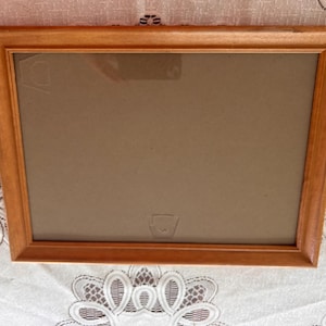 May include: Rectangular wooden picture frame with a light brown finish. The frame has a simple, classic design and a neutral-colored backing. The frame is empty, ready to display a photograph or artwork.