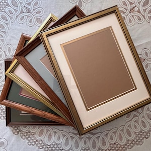May include: A collection of four rectangular picture frames stacked in a fan arrangement. The frames feature various finishes including metallic gold, dark wood grain, and medium brown wood, each equipped with off-white or colored matting.
