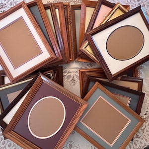 May include: A collection of rectangular wooden picture frames in various shades of brown, featuring white, cream, or colored matting with either rectangular or oval openings. The frames are arranged in a layered, overlapping cluster.