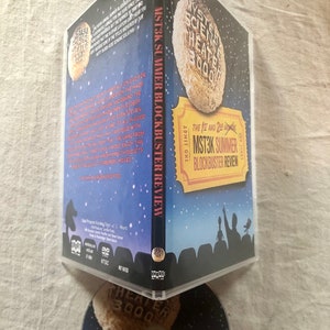 以下が含まれることがあります： 「ミステリー・サイエンス・シアター3000」の夏の話題作レビューのDVDケース。ケースは青色で、星でいっぱいの夜空の背景。前面には、「The 1st and 2nd Annual MST3K Summer Blockbuster Review」というテキストのチケットが描かれています。