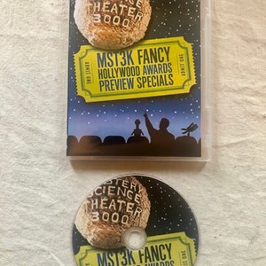 K&ouml;nnte beinhalten: Ein DVD-Geh&auml;use und eine Disc f&uuml;r die Mystery Science Theater 3000-Ver&ouml;ffentlichung "MST3K Fancy: Hollywood Awards Preview Specials". Das Geh&auml;use hat einen schwarzen Hintergrund mit einem gelben Ticket und einem Comic-Mond. Die Disc hat ein &auml;hnliches Design.
