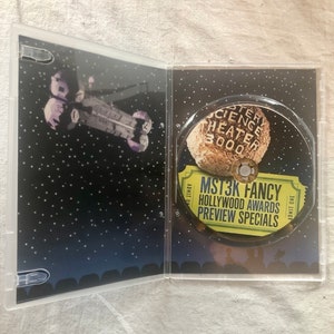 K&ouml;nnte beinhalten: Ein DVD-Geh&auml;use mit schwarzem Hintergrund und einem silbernen Raumschiff im Hintergrund. Die Vorderseite des Geh&auml;uses zeigt ein gelbes Ticket mit dem Text "MST3K FANCY HOLLYWOOD AWARDS PREVIEW SPECIALS" und ein Schwarzwei&szlig;bild eines Mondes mit dem Text "Mystery Science Theater 3000" darauf.