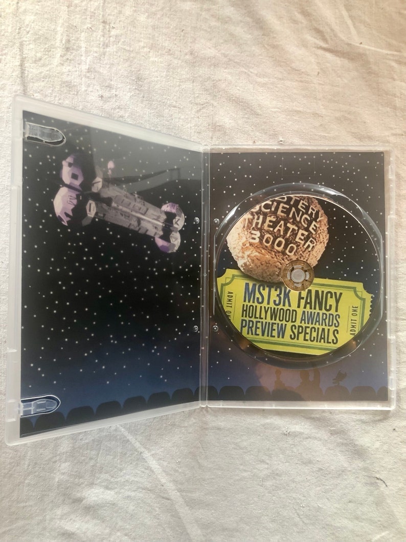 K&ouml;nnte beinhalten: Ein DVD-Geh&auml;use mit schwarzem Hintergrund und einem silbernen Raumschiff im Hintergrund. Die Vorderseite des Geh&auml;uses zeigt ein gelbes Ticket mit dem Text "MST3K FANCY HOLLYWOOD AWARDS PREVIEW SPECIALS" und ein Schwarzwei&szlig;bild eines Mondes mit dem Text "Mystery Science Theater 3000" darauf.