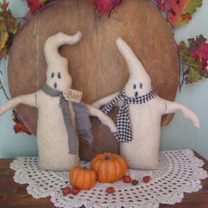 May include: Two white felt ghost figures with black and white checkered scarves, one with a tag that says "Boo!"  The ghosts are sitting on a white crocheted doily with two orange pumpkins and red berries.