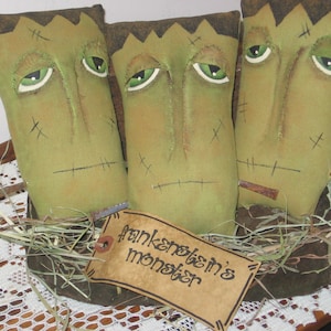 May include: Three green felt Frankenstein monster dolls with stitched details and painted faces. The dolls are sitting on a brown surface with a brown and green fabric base and a lace doily. A brown paper tag with the text "Frankenstein's monster" is in front of the dolls.
