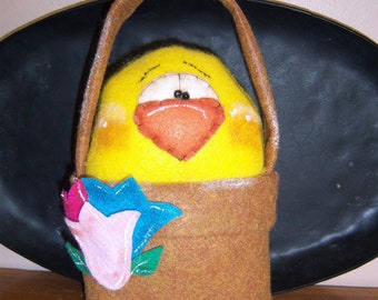 May include: A yellow felt chick with a red felt beak and black eyes is sitting in a brown felt bag with a pink and blue felt flower. The bag has a brown felt handle.