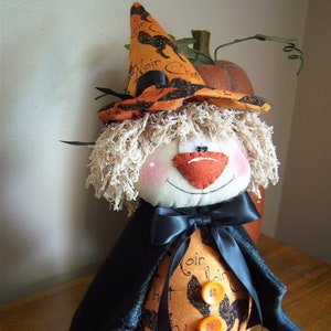 May include: A handmade Halloween witch doll with a black hat, a black cape, and orange and black fabric. The doll has a white face with pink cheeks and a red nose. The doll is sitting on a wooden surface.