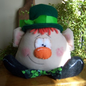 Puede incluir: Un juguete de peluche con forma de duende que lleva un sombrero verde con una banda verde y blanca. El juguete tiene barba roja, nariz naranja y una gran sonrisa.