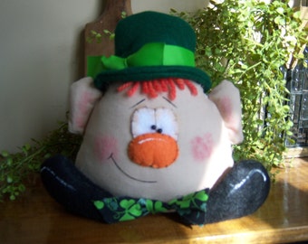 Pode incluir: Um brinquedo de pelúcia em forma de duende usando um chapéu verde com uma faixa verde e branca. O brinquedo tem barba vermelha, nariz laranja e um grande sorriso.