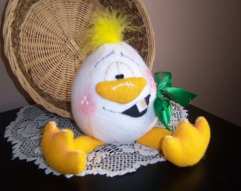 Pode incluir: Um brinquedo de pelúcia branco em forma de ovo com um bico amarelo, pés amarelos e um laço verde. O brinquedo tem um rosto pintado com um sorriso cheio de dentes e bochechas cor-de-rosa.