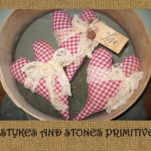May include: Three heart-shaped fabric ornaments with a red and white gingham pattern, adorned with cream-colored lace and tied with cream-colored ribbon. One heart has a tag that says "Love". The ornaments are displayed in a wooden sieve.