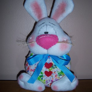 May include: A white plush bunny with pink ears and a blue bow. The bunny is wearing a colorful, splatter-painted outfit with two red hearts.