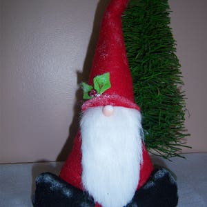 May include: A red felt gnome with a white beard and black boots sits in front of a green artificial Christmas tree. The gnome has a red felt hat with a white pom-pom and a green holly sprig.