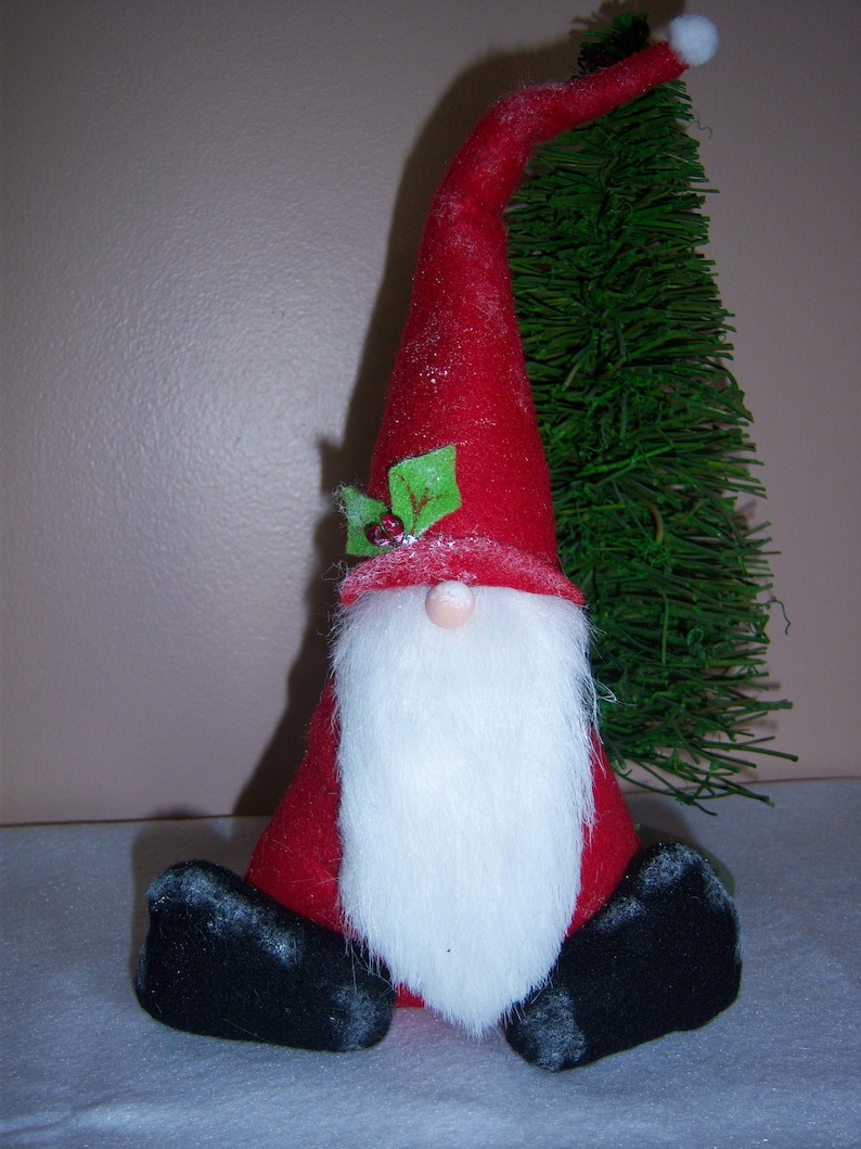 May include: A red felt gnome with a white beard and black boots sits in front of a green artificial Christmas tree. The gnome has a red felt hat with a white pom-pom and a green holly sprig.