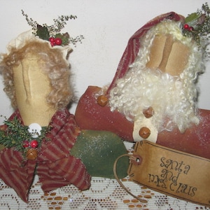 May include: A handmade Christmas decoration set featuring a Santa and Mrs. Claus doll. The dolls are made of fabric and have a rustic, vintage look. The Santa doll is wearing a red and white striped hat and a white beard. The Mrs. Claus doll is wearing a red and white striped scarf. The dolls are sitting on a green and white fabric base. The set includes a brown paper tag with the text "Santa and Mrs. Claus".